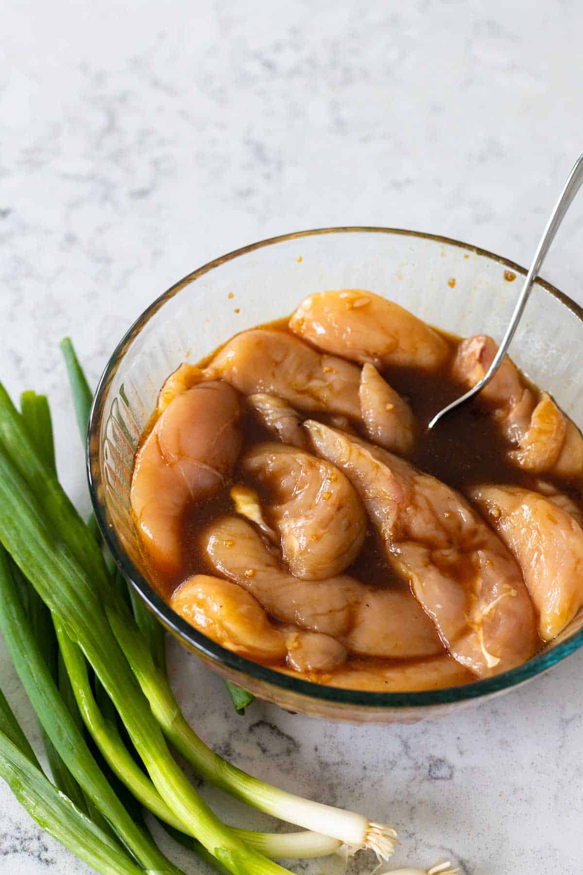 A mixing bowl is filled with chicken tenderloins marinating in the Asian marinade. There's a bunch of green onions on the side.