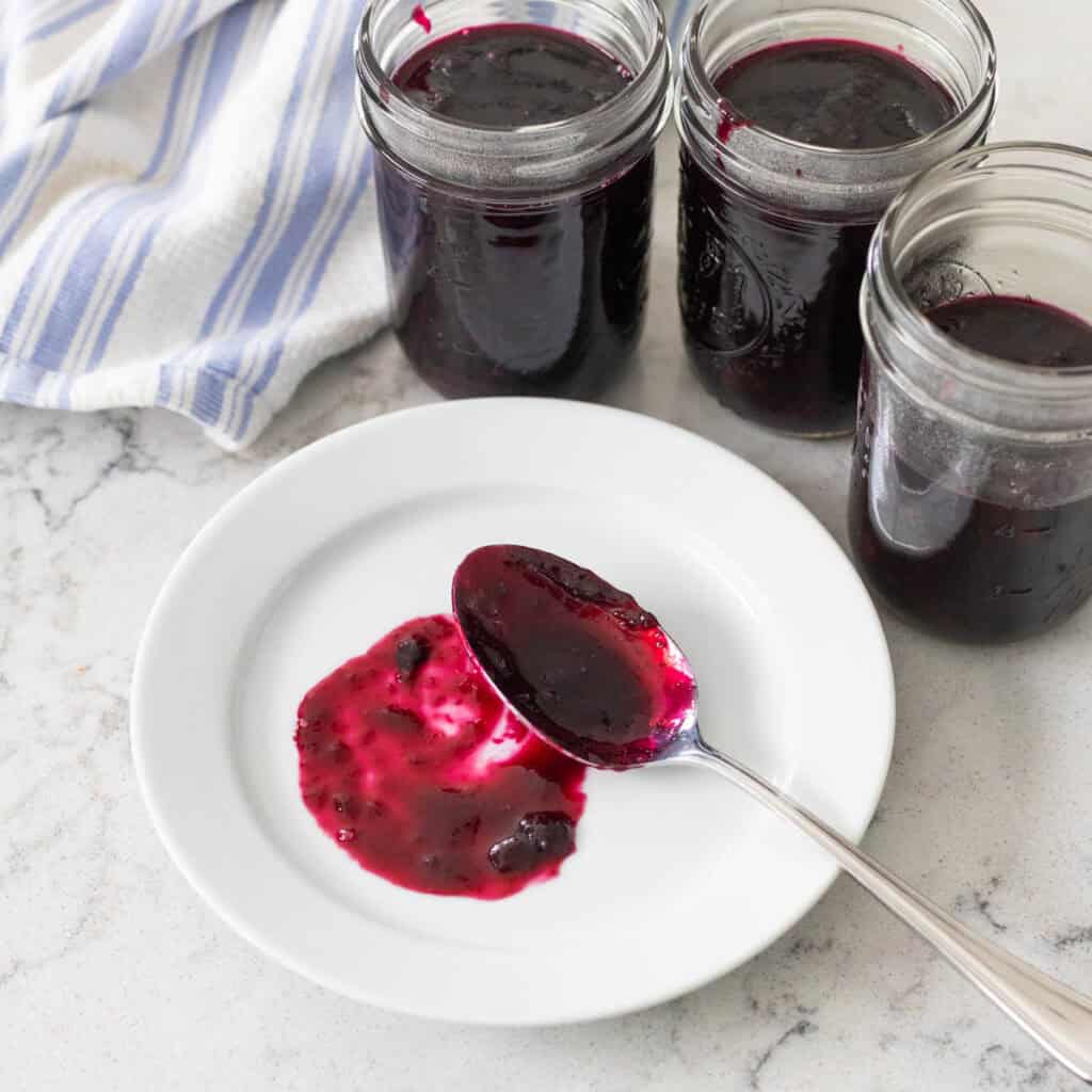3 mason jars filled with blueberry preserves surround a white plate with a spoon filled with the jam spread to show the color.