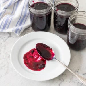 3 mason jars filled with blueberry preserves surround a white plate with a spoon filled with the jam spread to show the color.