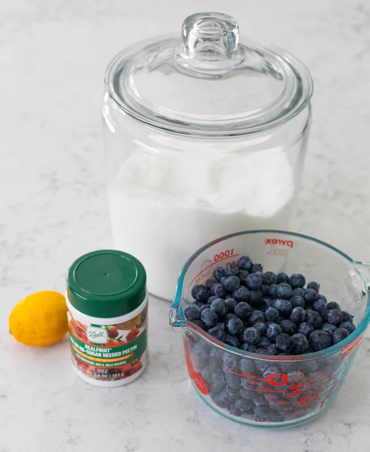 Fresh blueberries sit next to a jar of sugar and a container of pectin. The lemon is off to the side.