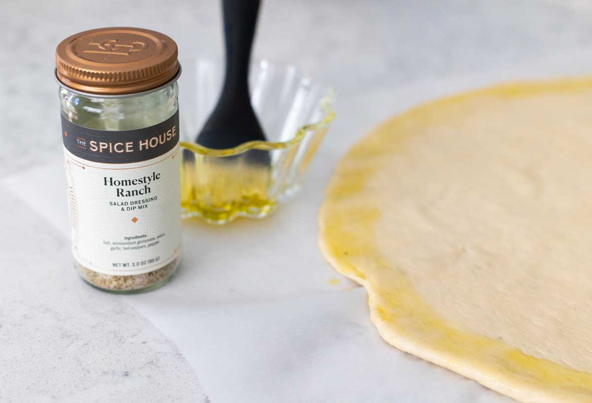 A jar of spices next to a bowl of olive oil and a pizza dough that has been brushed with the oil.