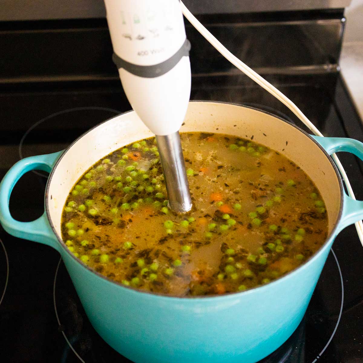 An immersion blender is pureeing the soup. Seasonings are floating on the top of the broth.