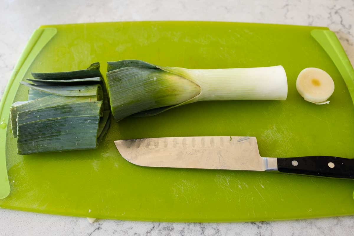 The leek is on a cutting board. The top greens have been cut and the bottom roots sliced by a large chef knife.