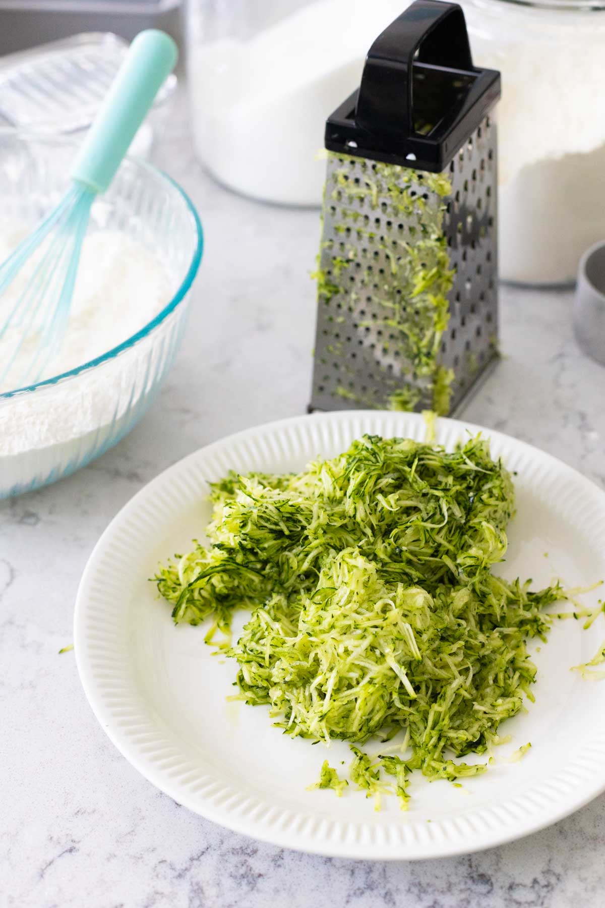 A white plate with grated zucchini has a box grater in the background.