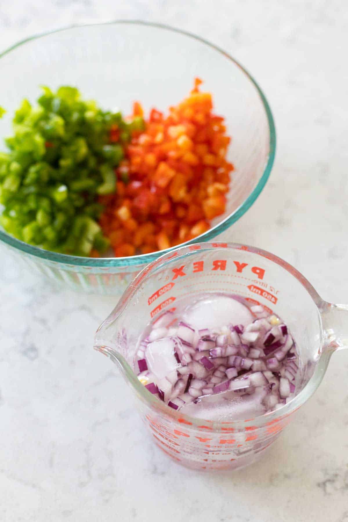 The red onions are soaking in an ice water bath. The chopped green and red bell peppers are in a bowl next to them.