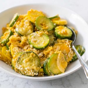 A white bowl is filled with sliced zucchini squash covered in cheddar cheese and bread crumbs.
