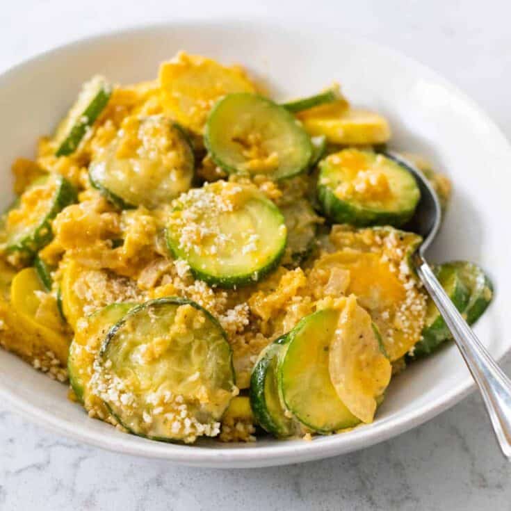 A white bowl is filled with sliced zucchini squash covered in cheddar cheese and bread crumbs.