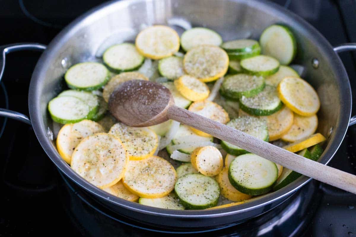 The squash have been added to the skillet and seasoned. A spoon is stirring them.