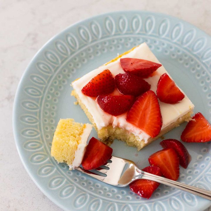 A square of almond cake with white almond icing is on a blue plate and topped with fresh strawberries. A fork is taking a bite.