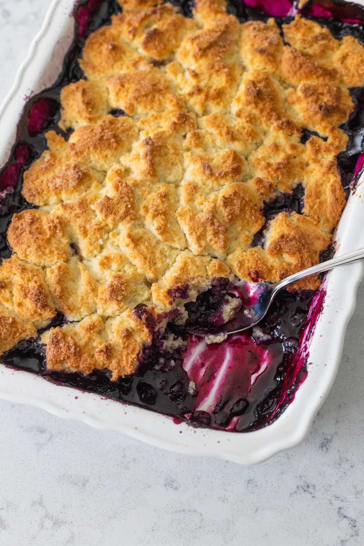 The finished cobbler has a spoon scraping out a serving. You can see the rich purple blueberry filling hiding under the golden brown topping.