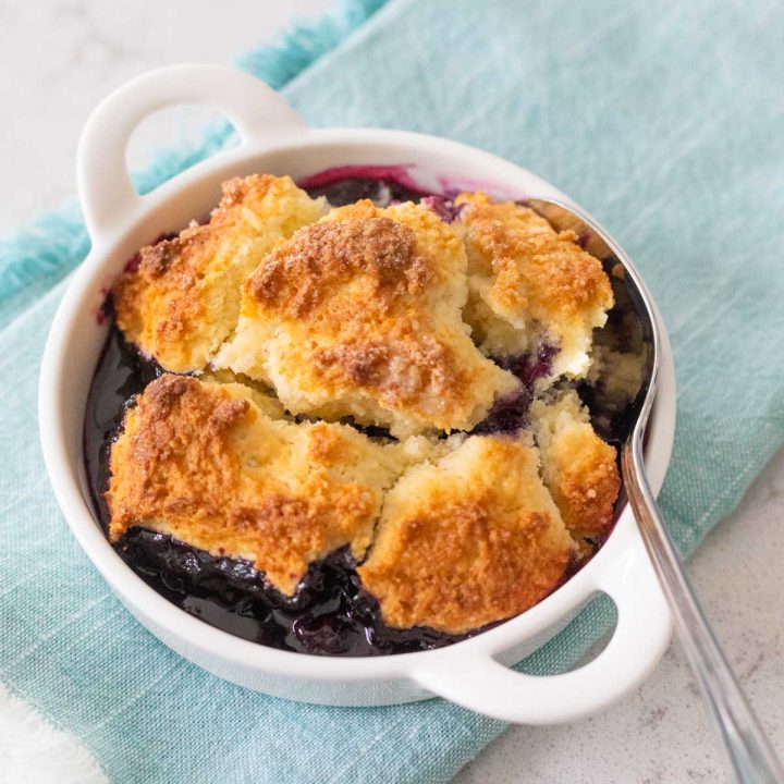 A white dish has the blueberry cobbler with golden brown topping ready to eat. A spoon is about to dig in.