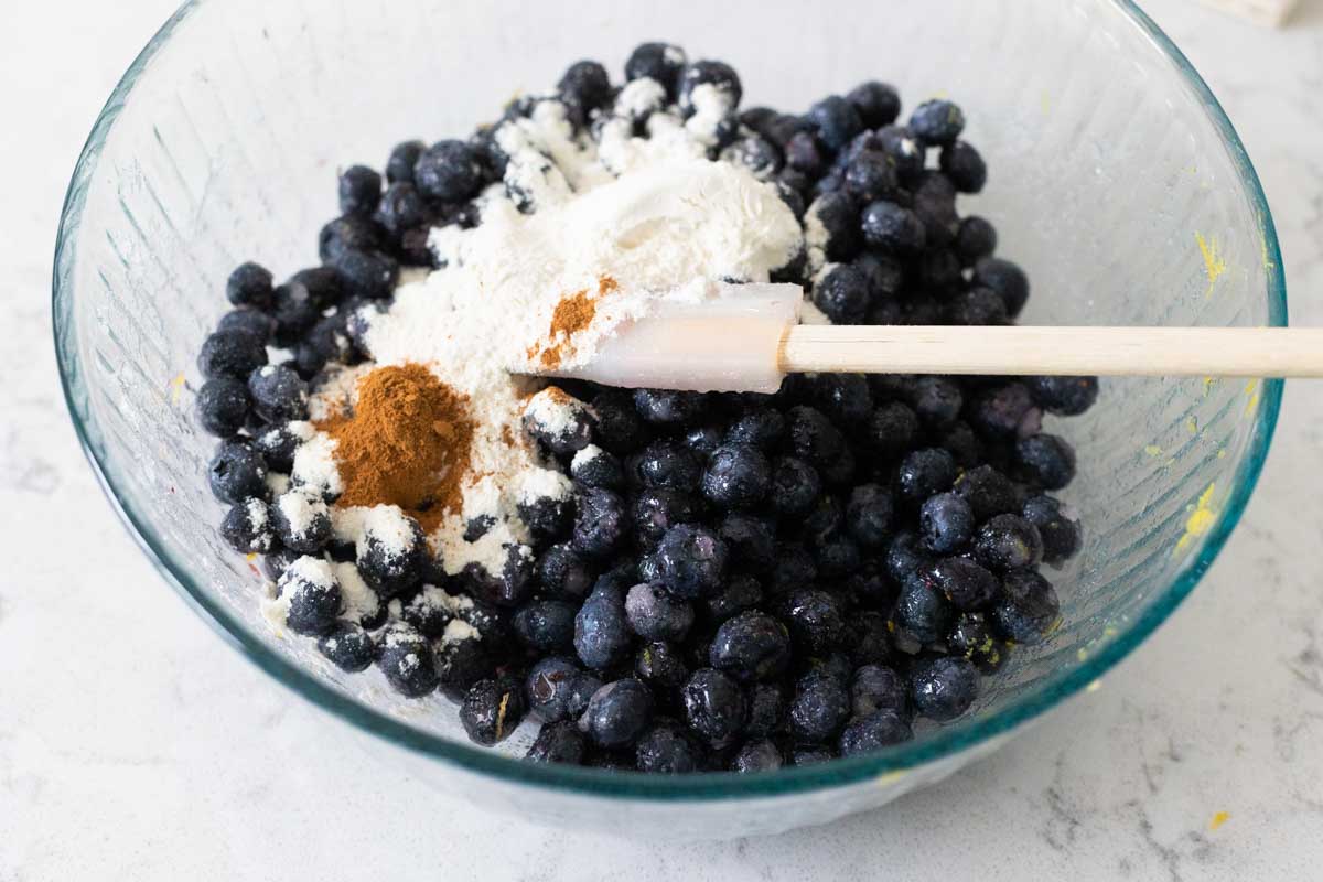 The flour and cinnamon have been added to the mixing bowl with blueberries.