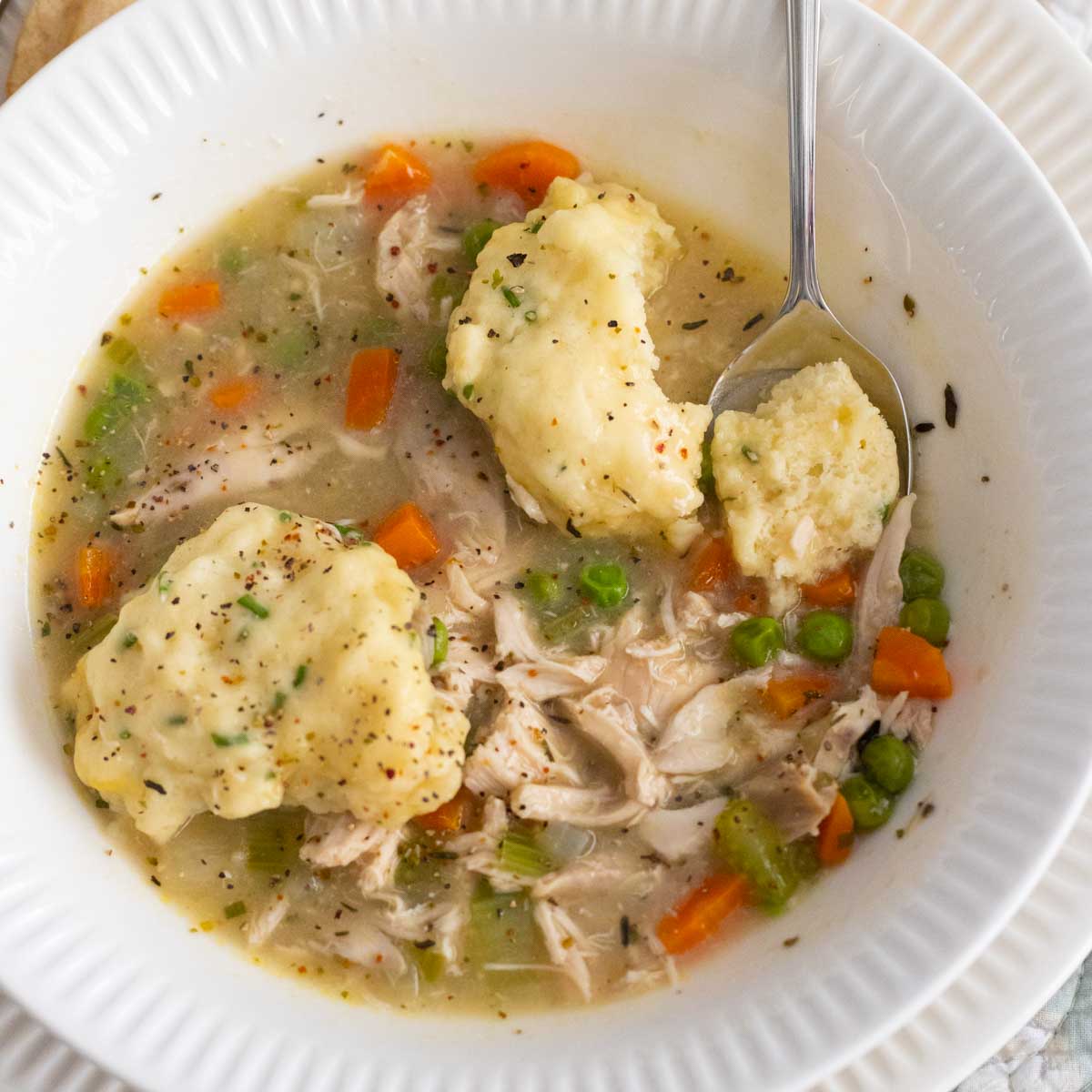 The white bowl has a serving of chicken and dumplings with a spoon taking a bite of the dumpling to show texture.