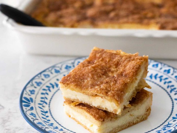 Two churro cheesecake bars are stacked on a blue and white plate in front of the baking dish of treats.