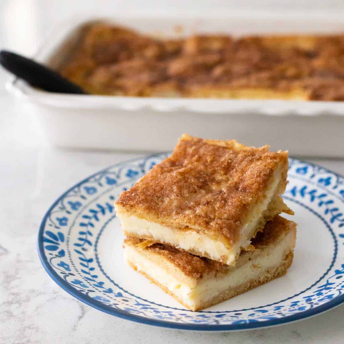 Two churro cheesecake bars are stacked on a blue and white plate in front of the baking dish of treats.