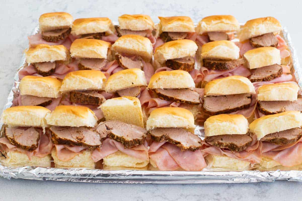 A tray of slider buns have been split and then filled with ham and a slice of pork tenderloin.