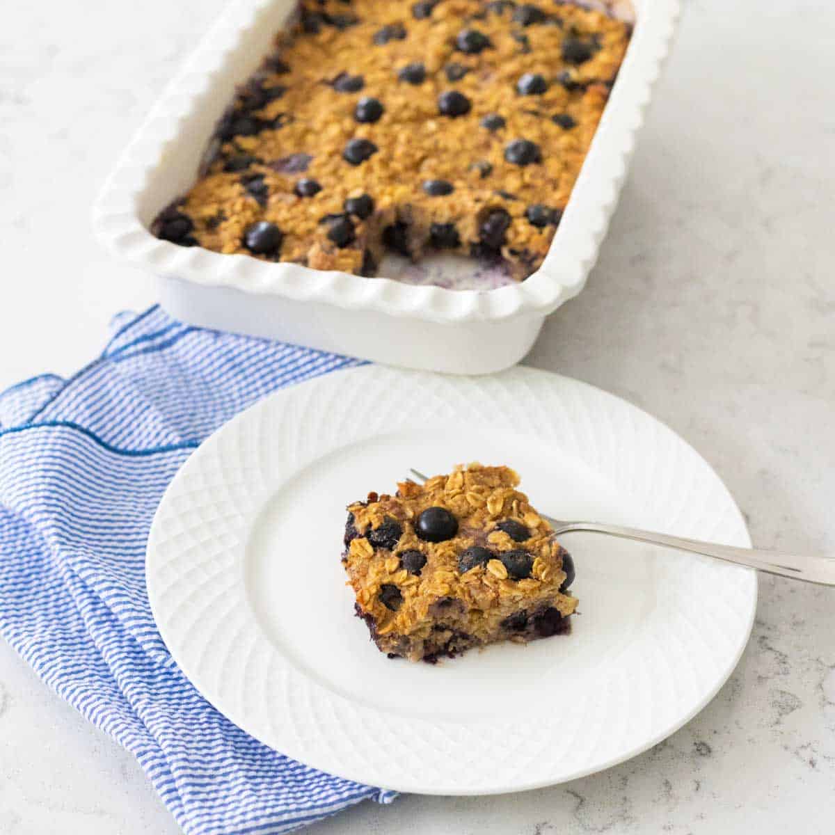 A square of baked oatmeal is on a white plate with a fork. There's a blue napkin on the side, the baking pan of oatmeal is in the background.