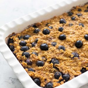A white baking dish is filled with the baked oatmeal. Blueberries are dotted over the top.