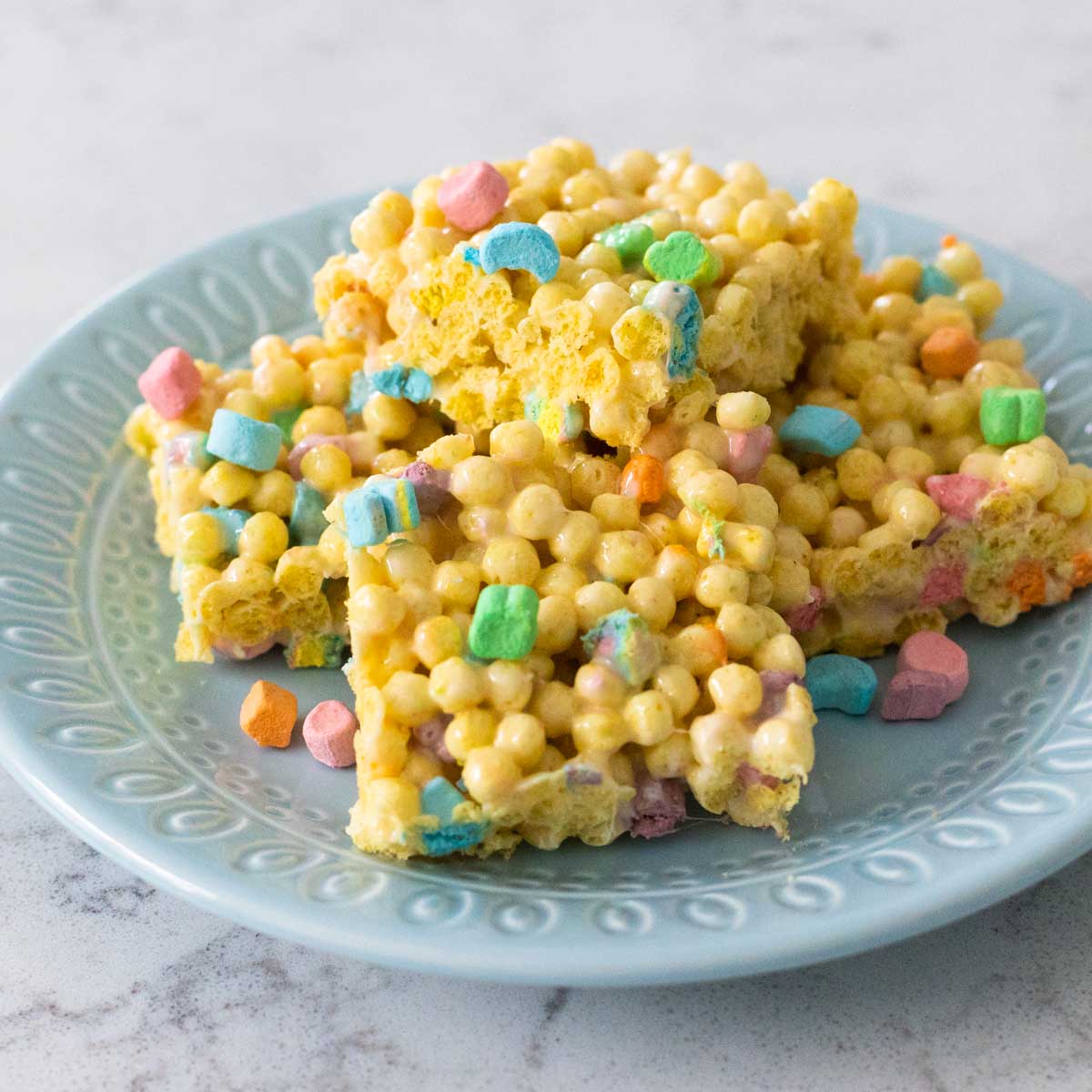 A blue plate has a pile of Mini Lucky Charms Krispie Treats with rainbow marshmallows.