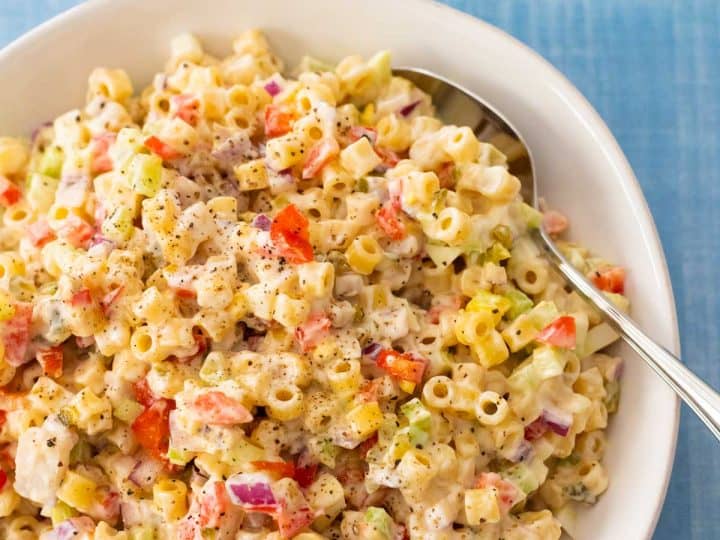 A white bowl is filled with classic macaroni salad that has onions, red pepper, and celery peeking out of the dressing.