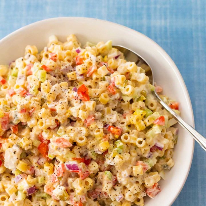 A white bowl is filled with classic macaroni salad that has onions, red pepper, and celery peeking out of the dressing.