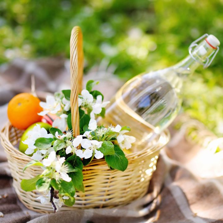 A wicker picnic basket with white flowers and a bottle of water with fresh fruit sits on a picnic blanket outside.