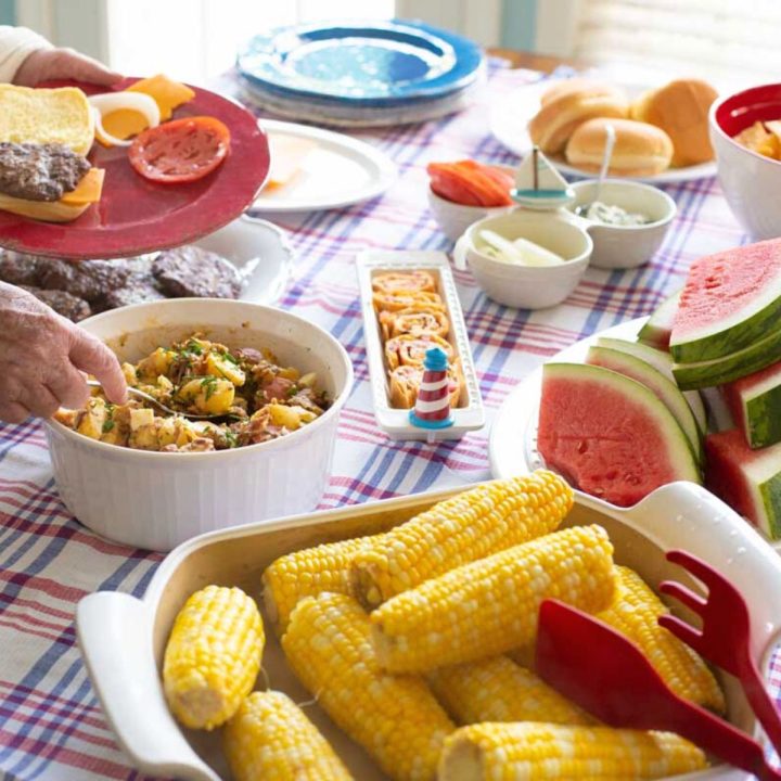 A potluck table is filled with 4th of July party recipes including fresh corn on the cob, watermelon, and potato salad.