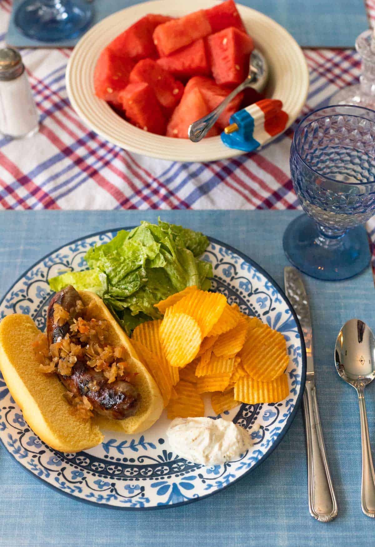 A blue and white plate has a grilled brat with potato chips and a green salad. A bowl of fresh watermelon is in the background.