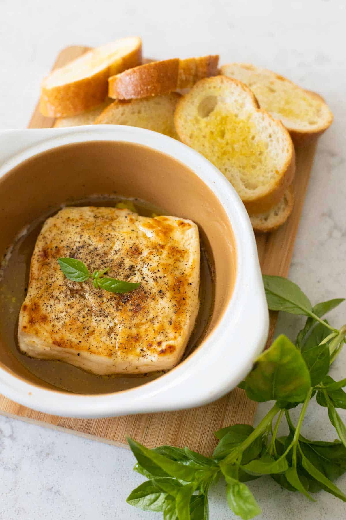 The baked feta is on a cutting board next to crostini. Black pepper and a sprig of basil are on top.