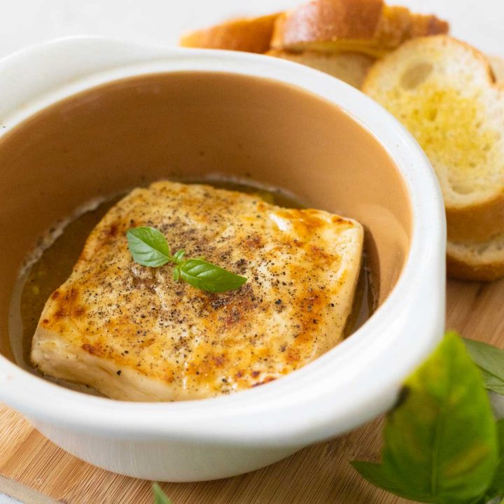 The white baking dish has a golden brown square of baked feta with a sprig of fresh basil on top.