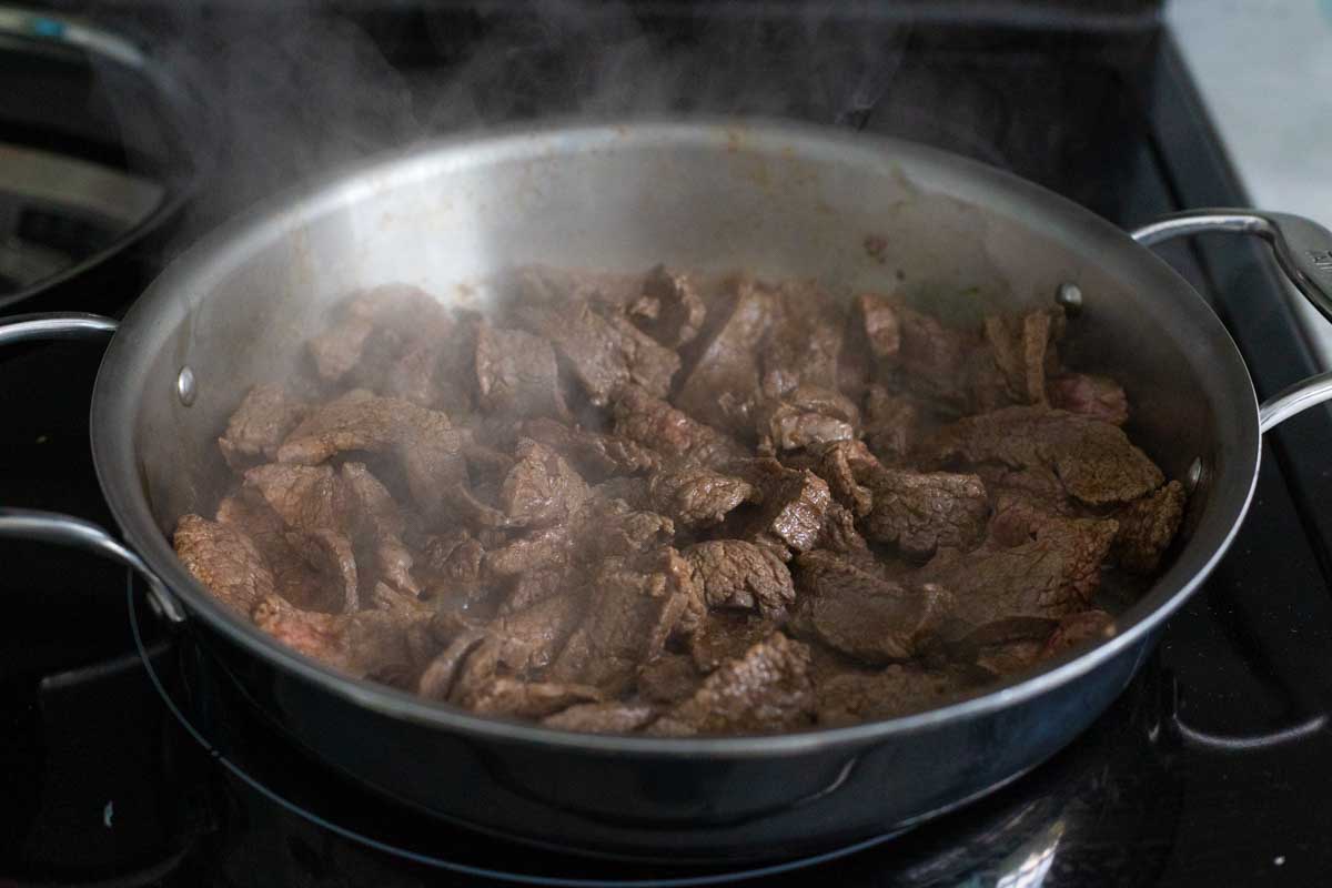 The strips of flank steak are being stir fried in a large skillet.