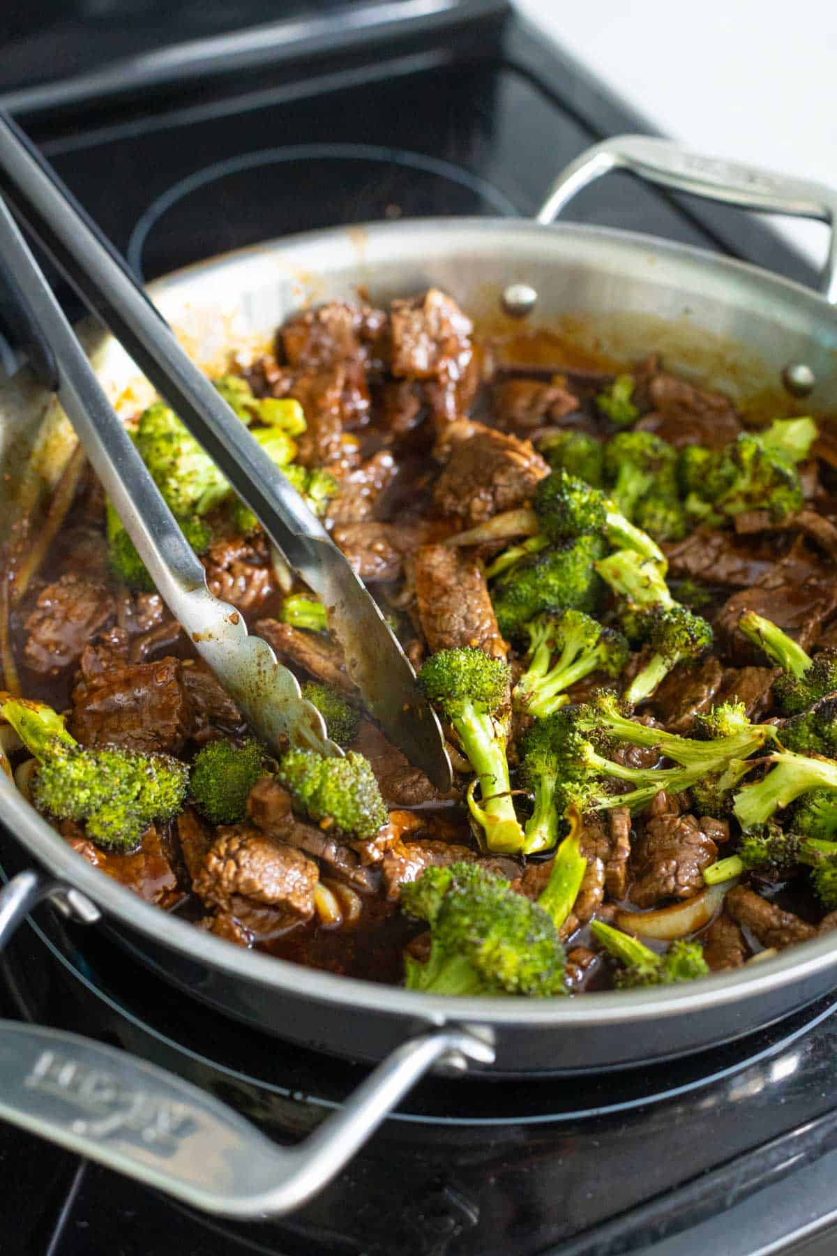 The finished beef and broccoli stir fry is in the skillet. A pair of tongs tosses everything in the sauce.