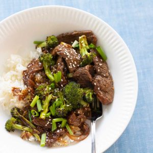 The white bowl has the stir fried beef and broccoli with chopped green onions over a bed of white rice against a blue background.