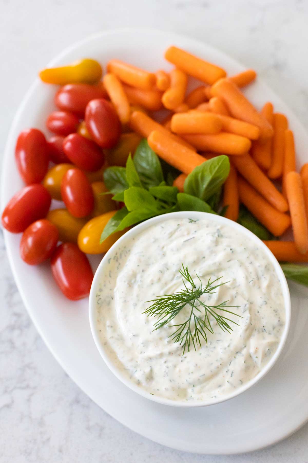 A bowl of dill dip has a sprig of dill on the top. The platter has cherry tomatoes, baby carrots, and a sprig of green basil for color.