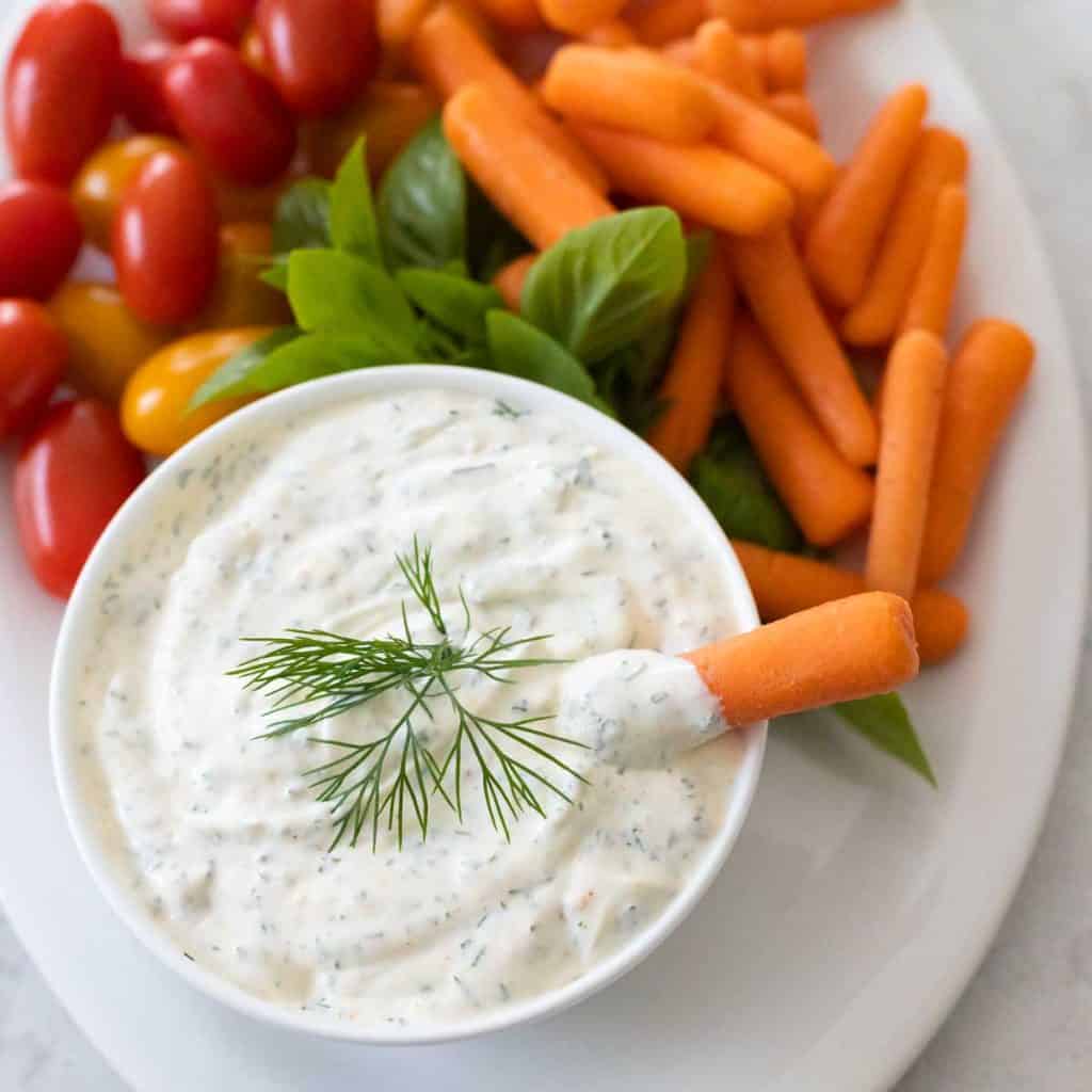 A bowl of dill dip has a baby carrot dipping in it. Cherry tomatoes and fresh herbs are on the platter behind it.