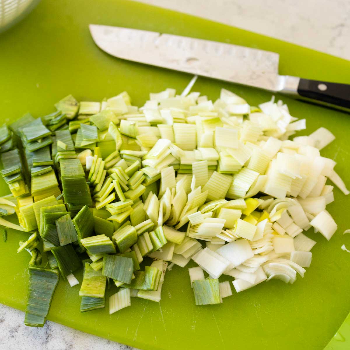A diced leek is on a green cutting board next to a large chef knife.
