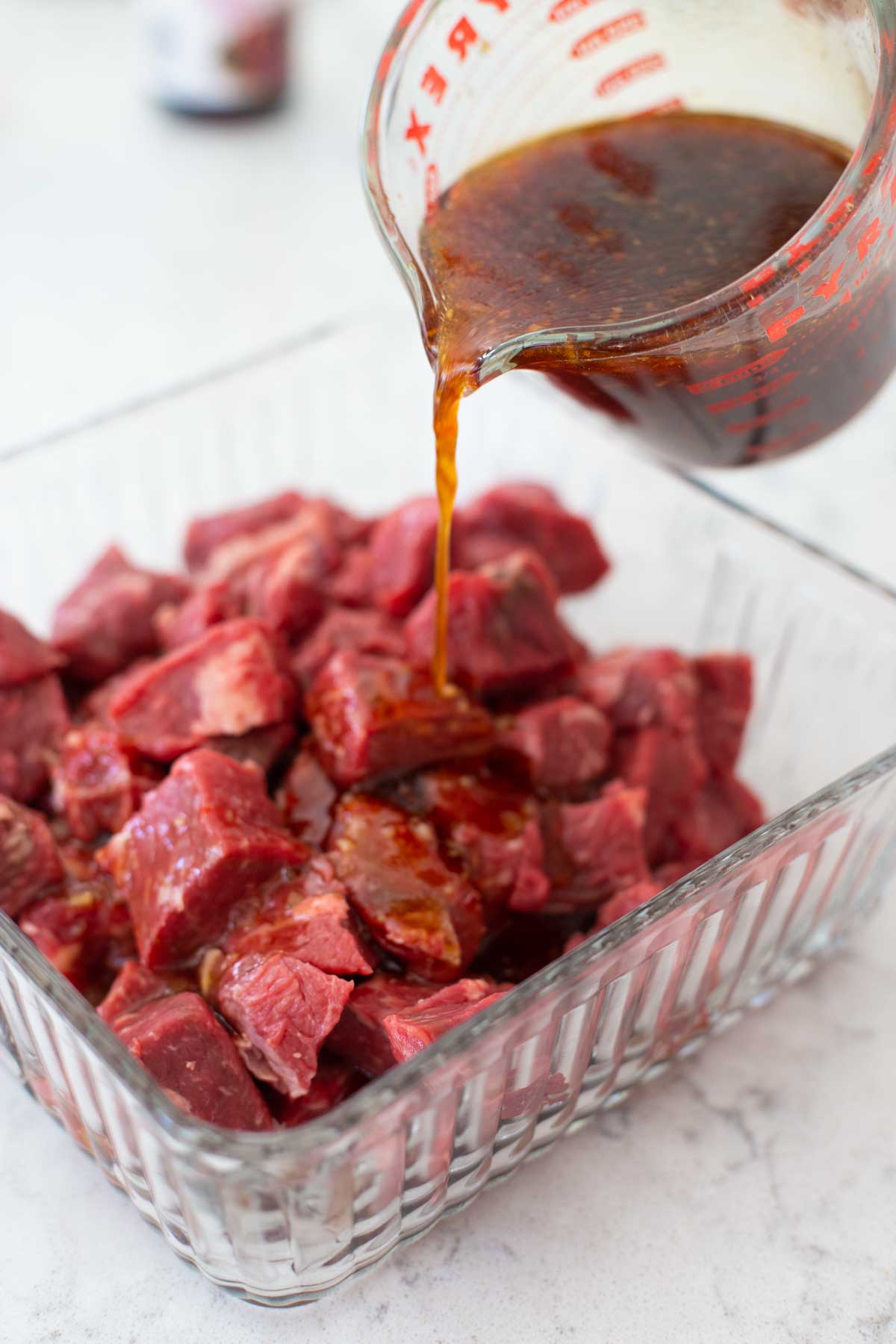 The chunks of beef are in a glass dish. The marinade is being poured over the top.