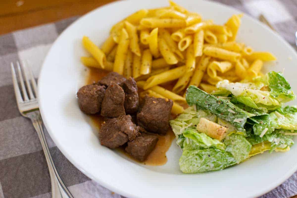 A white plate has the marinated steak next to a scoop of buttered penne pasta and a caesar salad.