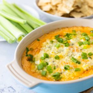 A baking dish filled with buffalo chicken dip sits next to a plate of celery and a bowl of tortilla chips.