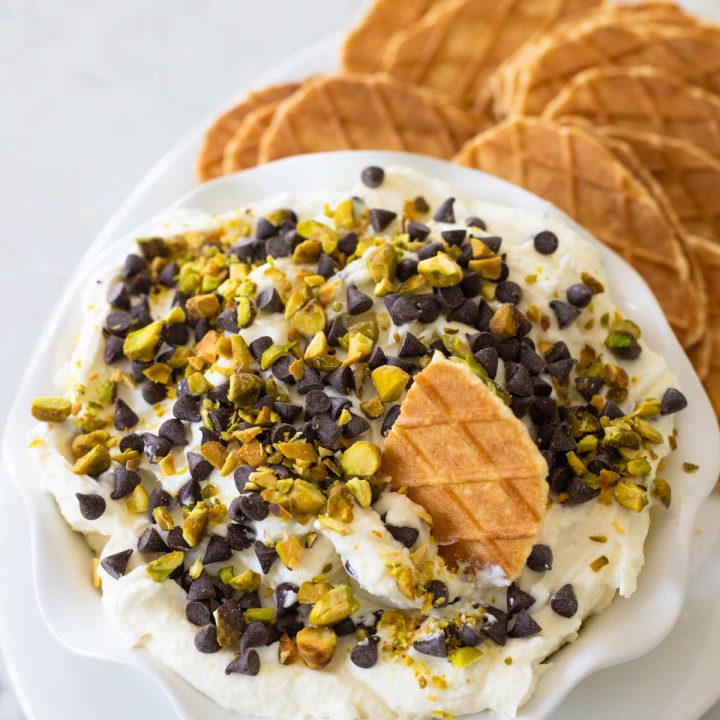 A ruffled bowl of cannoli dip has mini chocolate chips and chopped pistachios on top. A waffle cookie is being dunked into it.