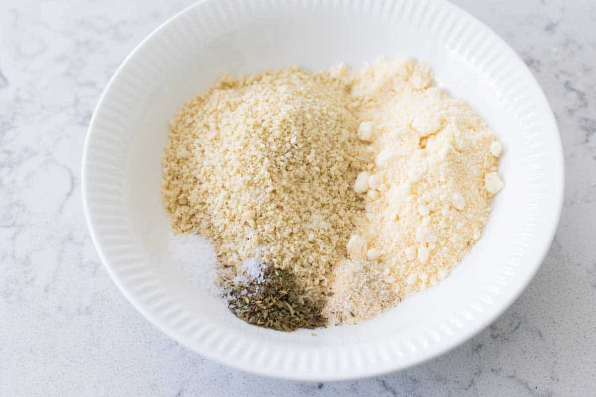 A white bowl has the bread crumbs, parmesan cheese, salt, and Italian herb blend.