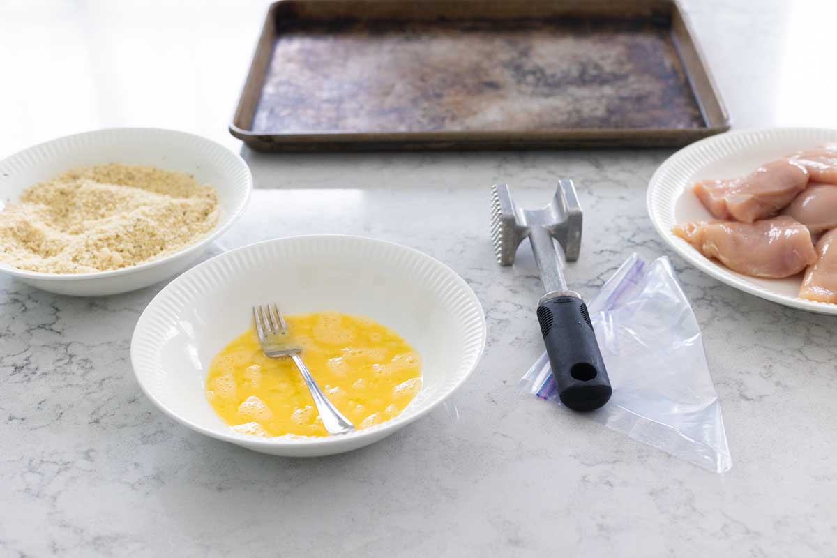 The chicken is on a plate on the right, the bowl of egg wash is in the middle, and the breadcrumbs are on the left. An empty baking sheet sits next to the station.