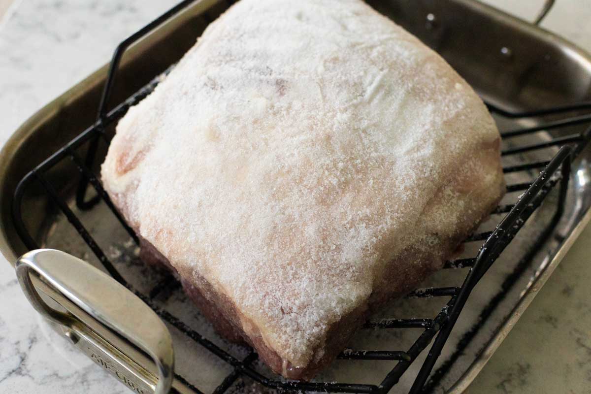 The pork shoulder sits in a roasting pan with a rack. It has been rubbed down and coated with lots of salt.