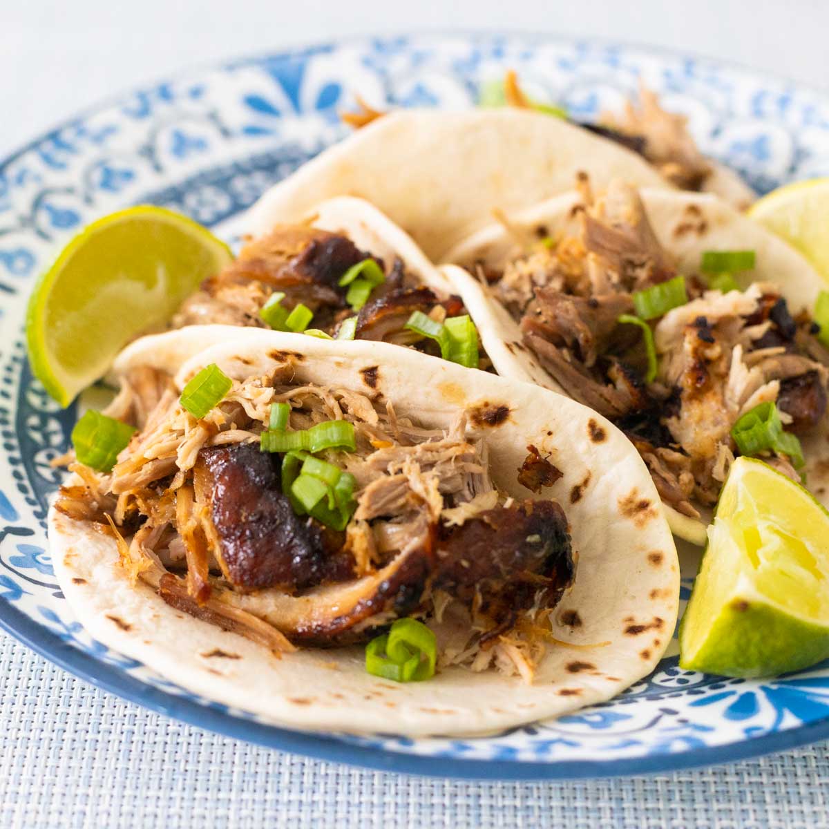 The shredded pork has been added to street taco tortilla shells. Green onions are sprinkled over the top and wedges of limes are on the side.