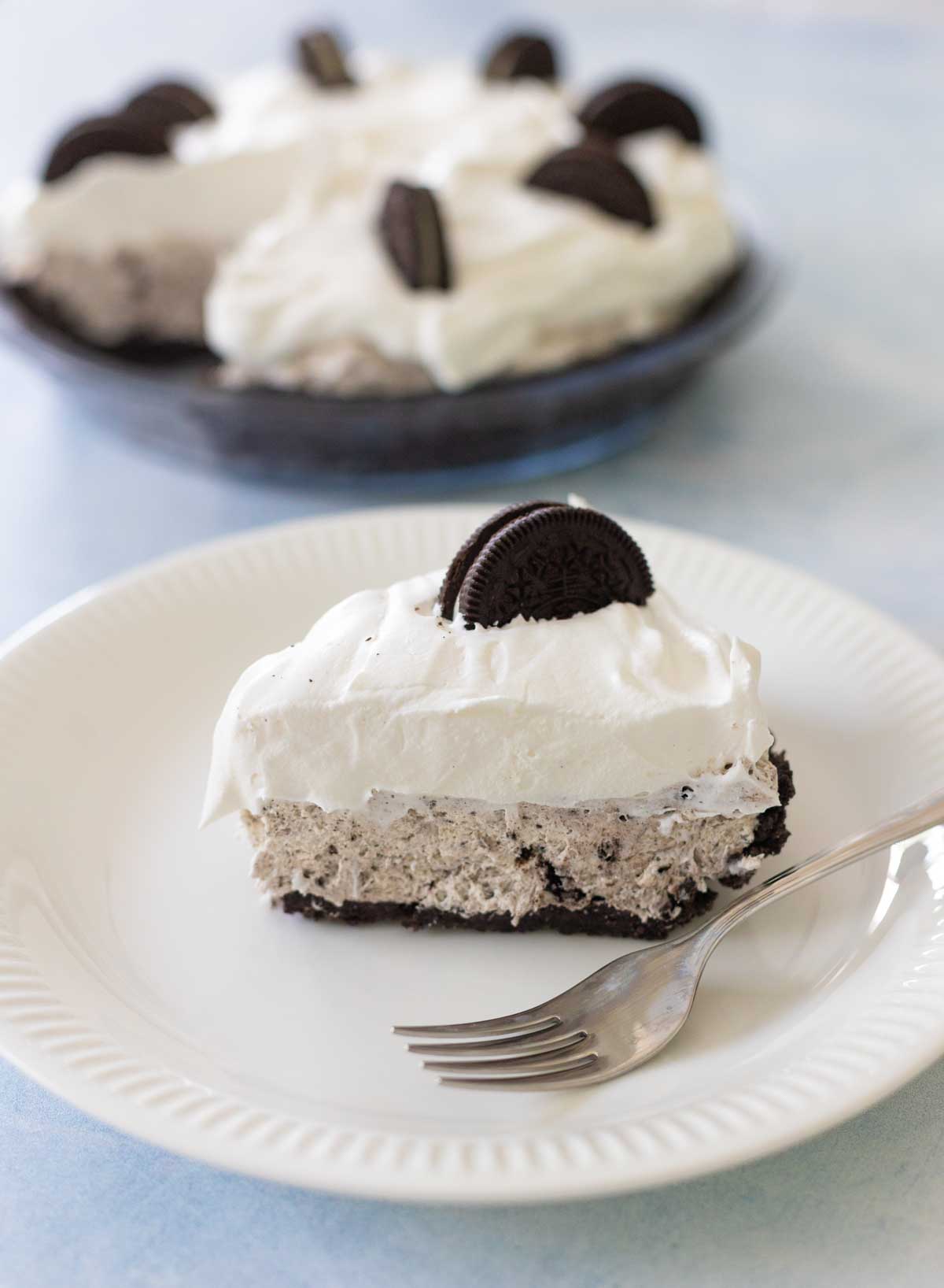 A slice of Oreo cream pie is on a white plate, the rest of the pie is in the background.