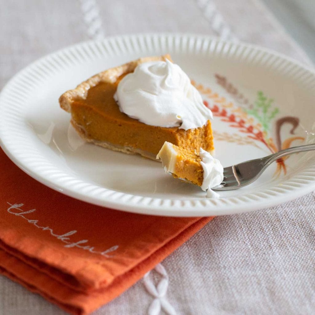 A slice of pumpkin pie on a fall harvest plate with an orange napkin.