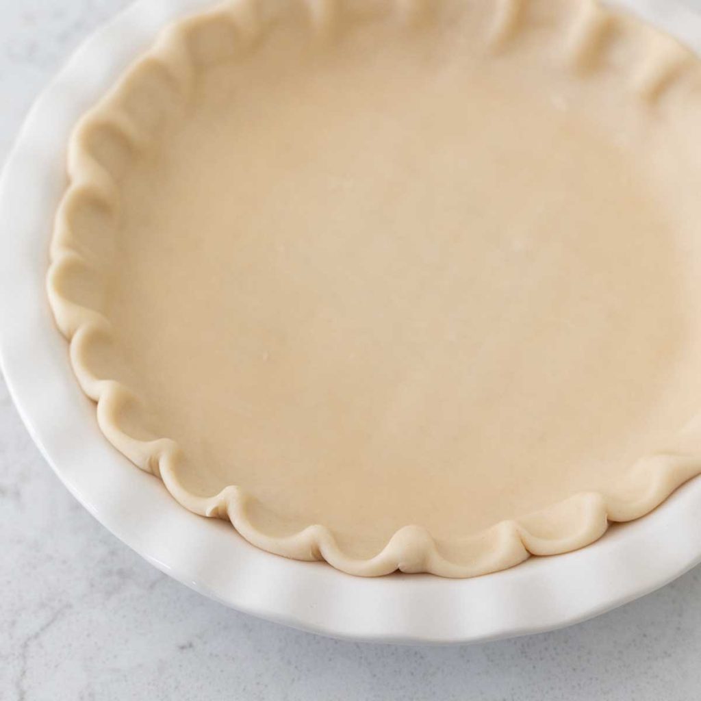 A white pie plate has a ruffled pie crust waiting to be filled with pie filling.