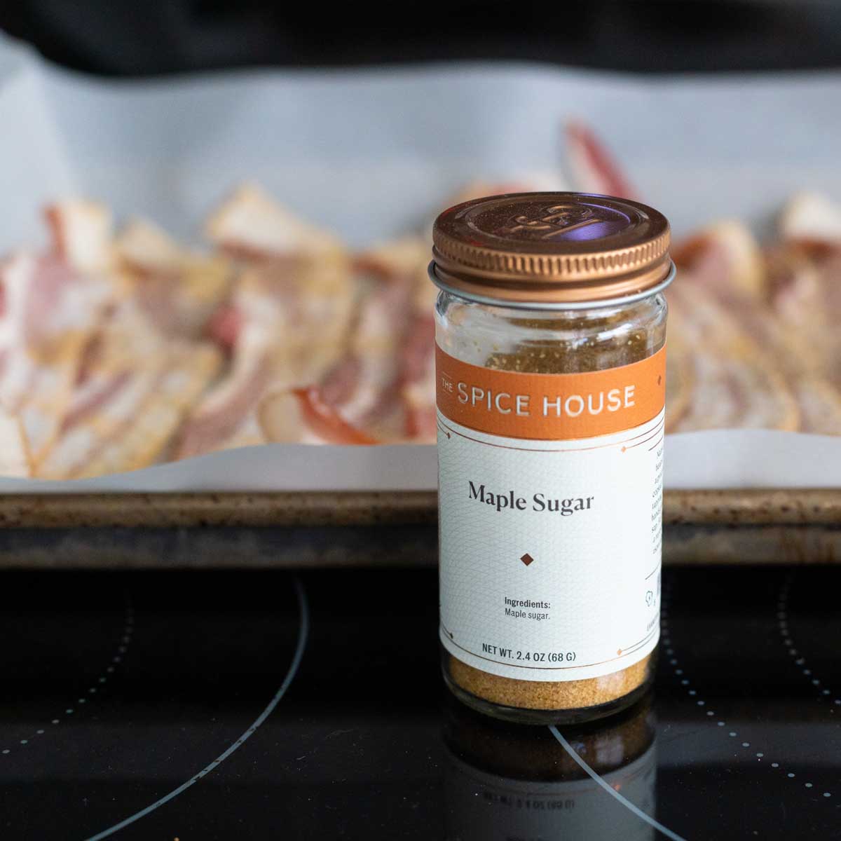 A jar of maple sugar sits in front of a pan of bacon.