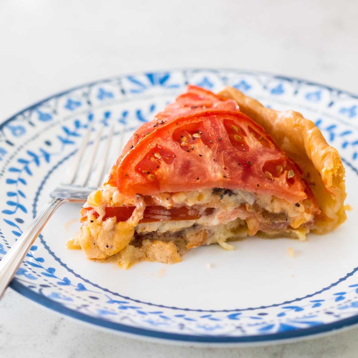 A slice of tomato pie is on a blue and white plate. You can see the layers of melted cheese and tomato slices.