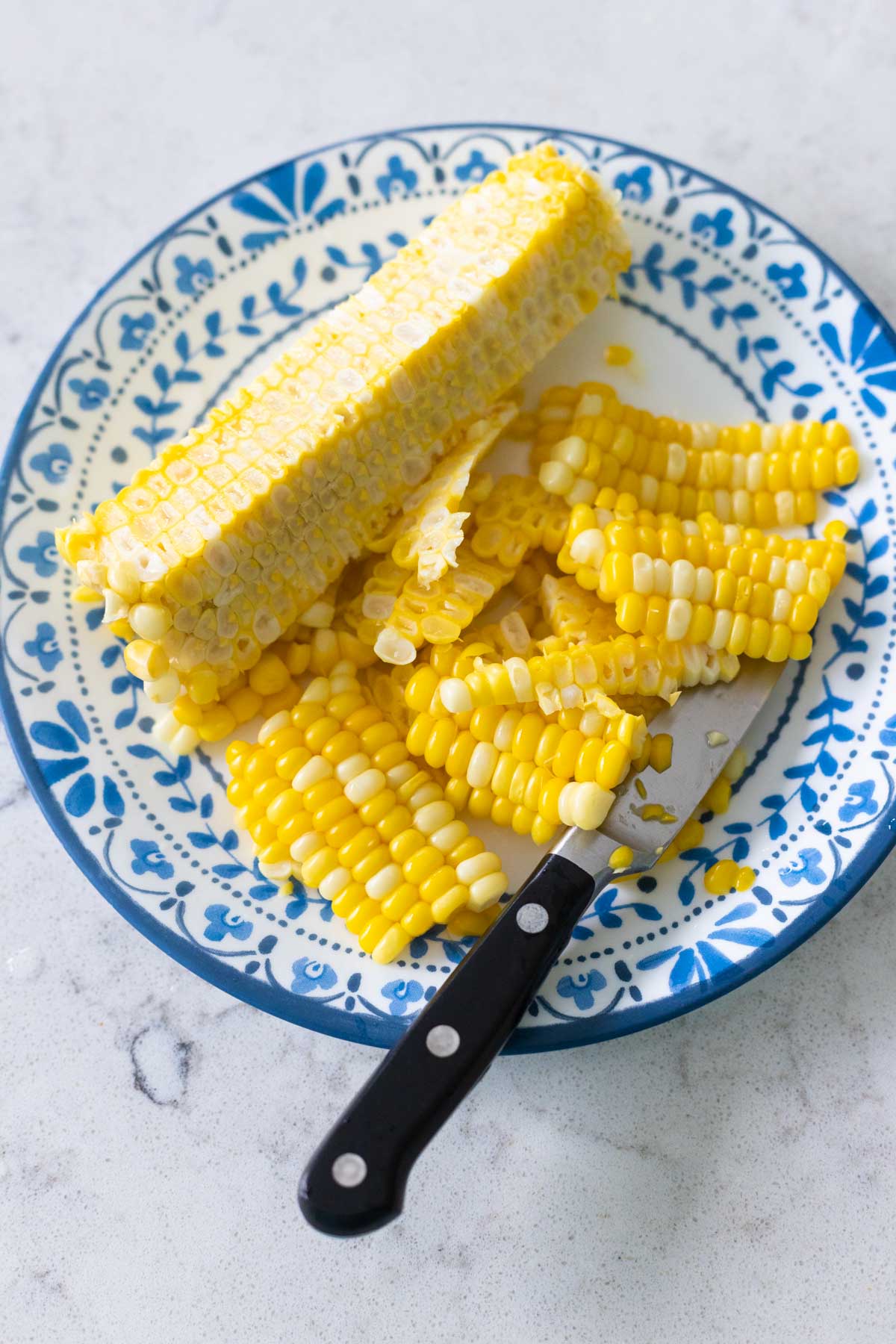 A cob of corn sits on a blue plate. A paring knife just removed all the corn kernels which rest on the plate next to the cob.